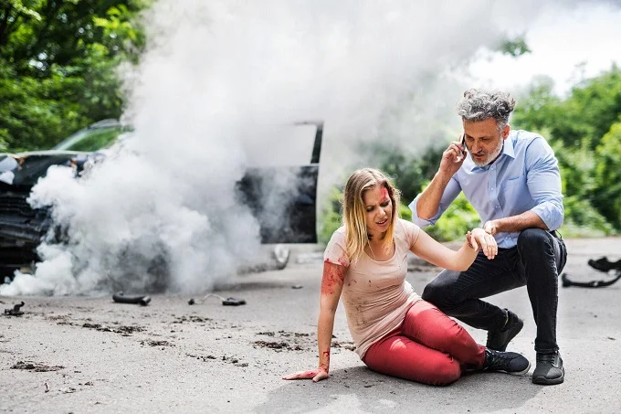A man assisting an injured woman on the roadside after a car accident, with a damaged car and smoke in the background.