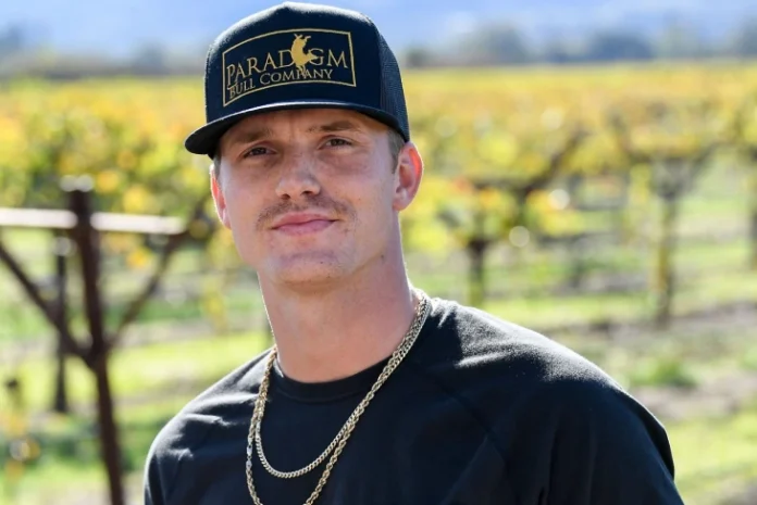Parker McCollum standing in a vineyard, wearing a Paradigm Hill Company cap and gold chains, representing Parker McCollum Net Worth and his success.