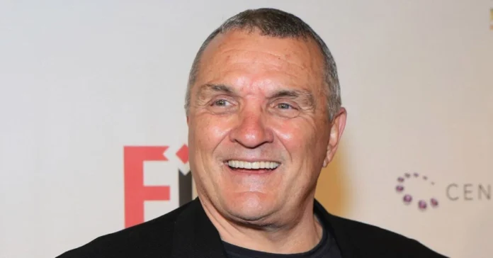 Rudy Ruettiger Net Worth Rudy Ruettiger smiling at an event, wearing a black jacket, representing his journey and success highlighted in the Rudy Ruettiger Net Worth article.