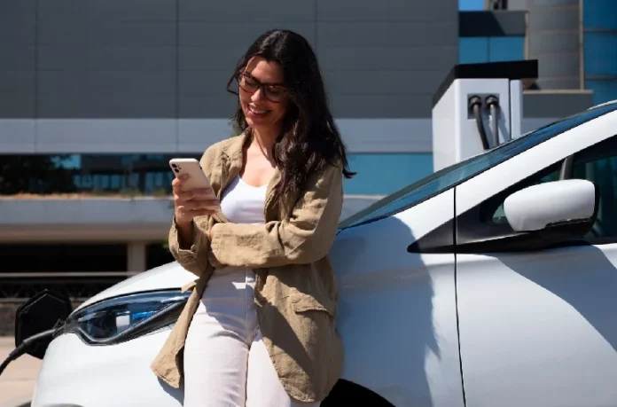 Owning an Electric Vehicle Woman leaning on an electric vehicle at a charging station, using her smartphone.